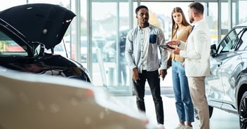 A salesperson inside a car showroom gesturing to a tablet while speaking with a young couple.