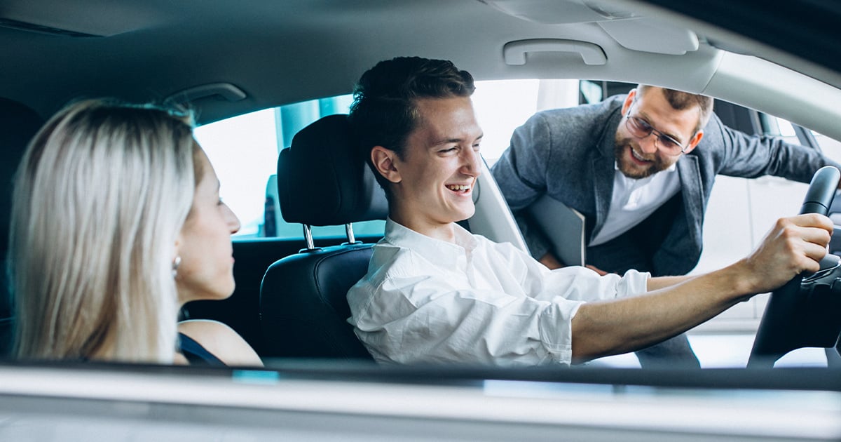 A young couple sitting in a new car inside a showroom laughing with the salesperson.