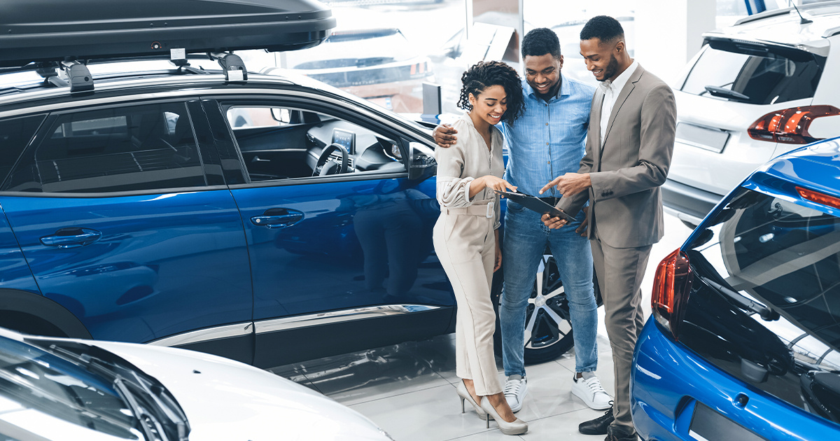 Couple Visiting Car Dealership Talking With Salesman In Auto Showroom