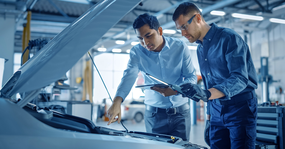 Two people in a auto dealership service department looking at the engine of a car while reviewing diagnostics on a tablet.